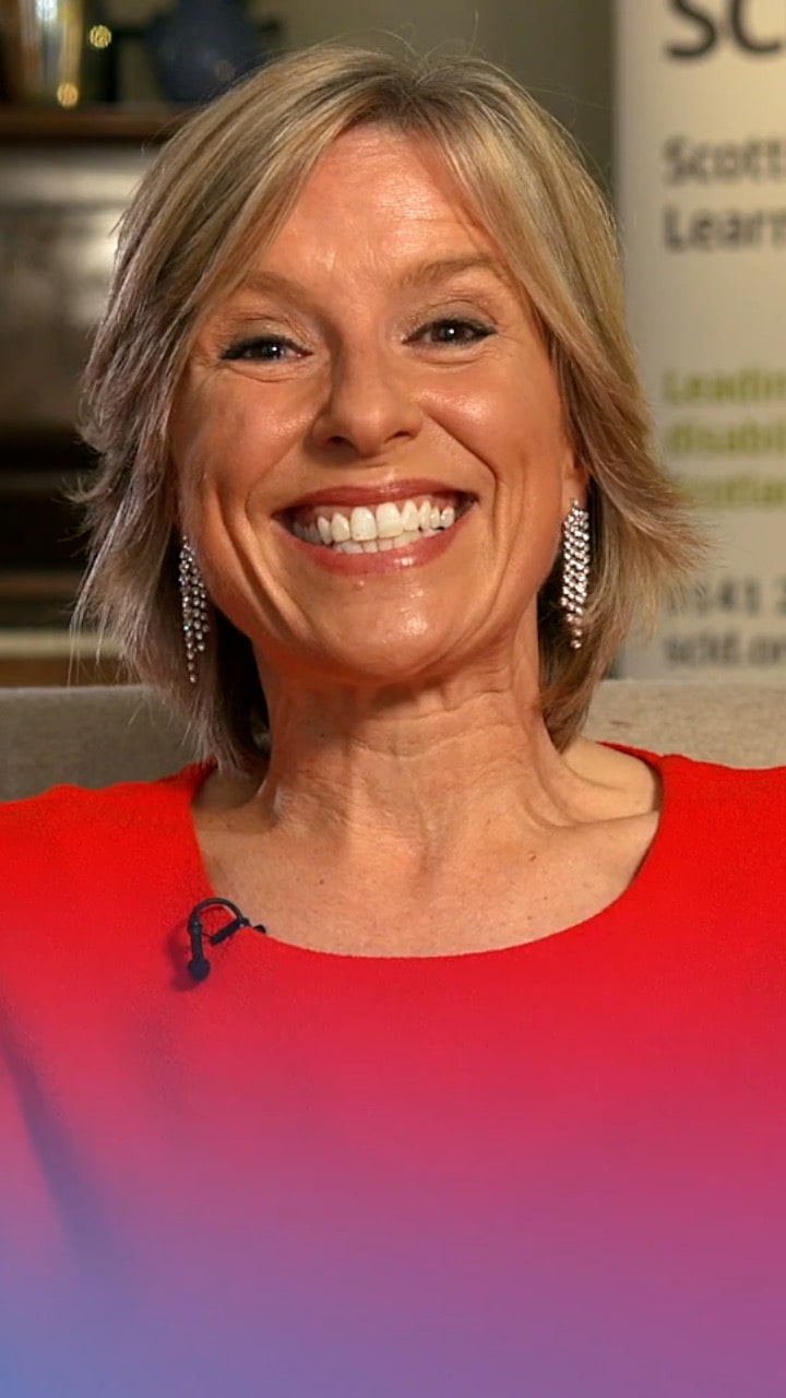 An image of a woman with short brown/blonde hair smiling at the camera. She has silver earrings hanging down from her ears and is wearing a red dress