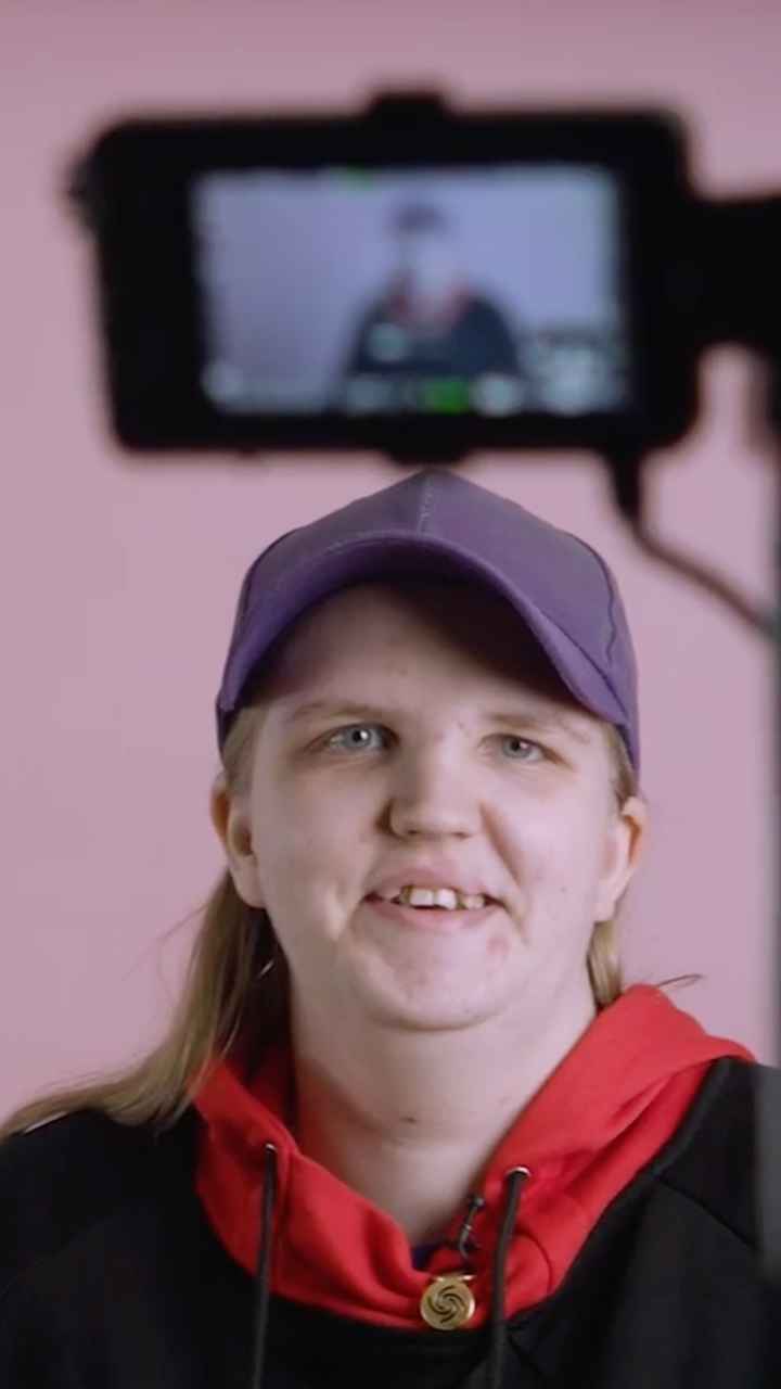 A young woman wearing a hooded top and baseball cap stares towards a camera screen in front of a pink background.